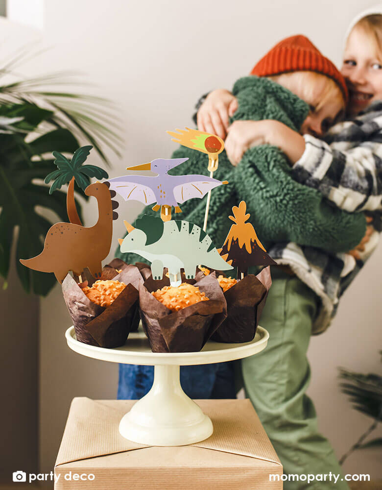 Two boys celebrating and holding each other each other at a dinosaur themed birthday party, in front of them are a few muffins topped with Momopartys's dinosaur themed cupcake toppers featuring dinosaurs, a volcano and a palm tree, setting a great prehistorical theme to a kid's dinosaur birthday party.