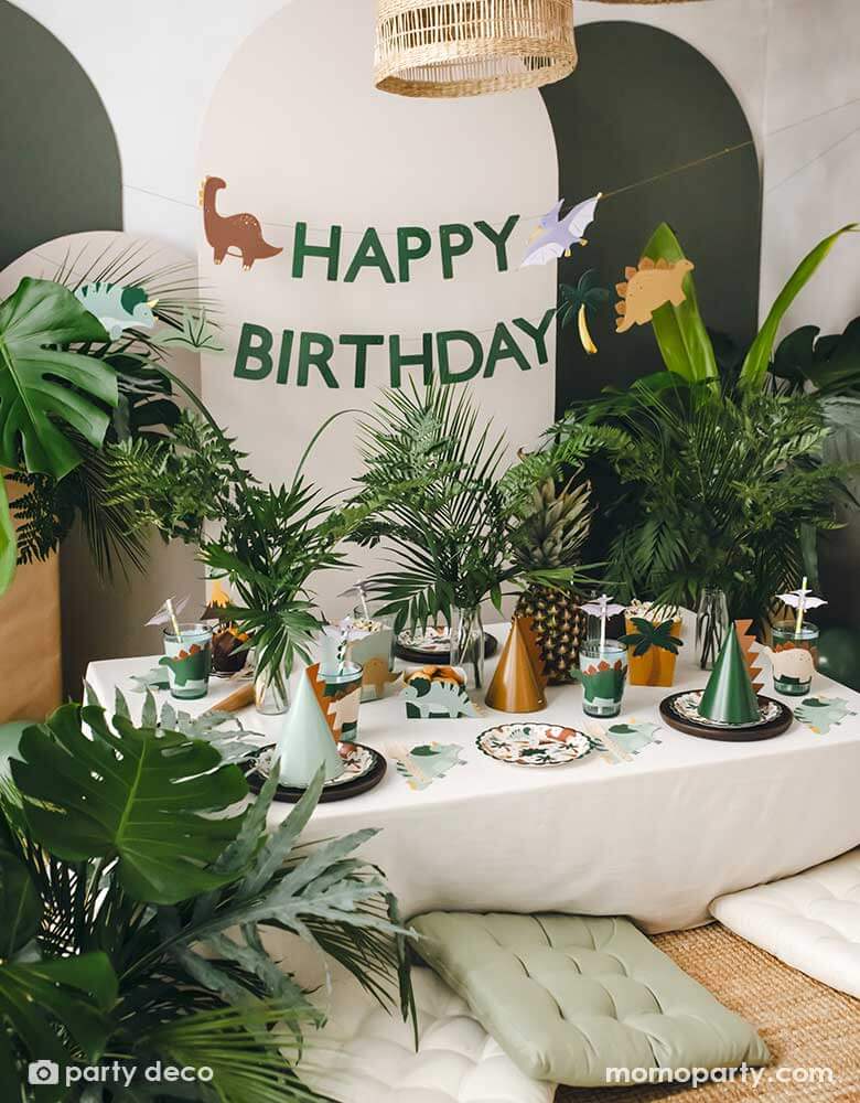 An adorable kid's dinosaur themed party set up featuring Momopartys's dinosaur themed party supplies and decorations by Party Deco including dinosaur paper plates, cup sleeves, dinosaur cake toppers, dinosaur spikes party hats, dinosaur napkins and a dinosaur happy birthday banner hung on the party backdrop wall. With a lot of tropical plants as the centerpiece, it makes a cute and modern inspo for kid's dinosaur themed birthday party.