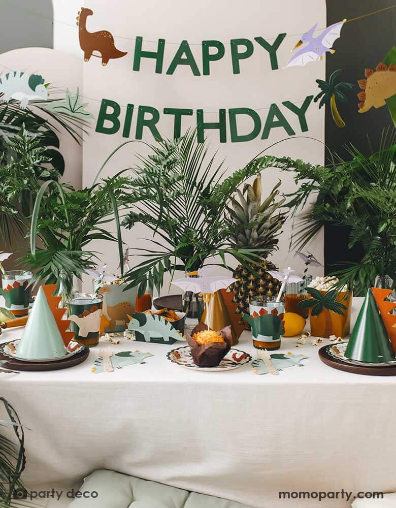 An adorable kid's dinosaur themed party set up featuring Momopartys's dinosaur themed party supplies and decorations by Party Deco including dinosaur paper plates, cup sleeves, dinosaur cake toppers, dinosaur spikes party hats, dinosaur napkins and a dinosaur happy birthday banner hung on the party backdrop wall. With a lot of tropical plants as the centerpiece, it makes a cute and modern inspo for kid's dinosaur themed birthday party.
