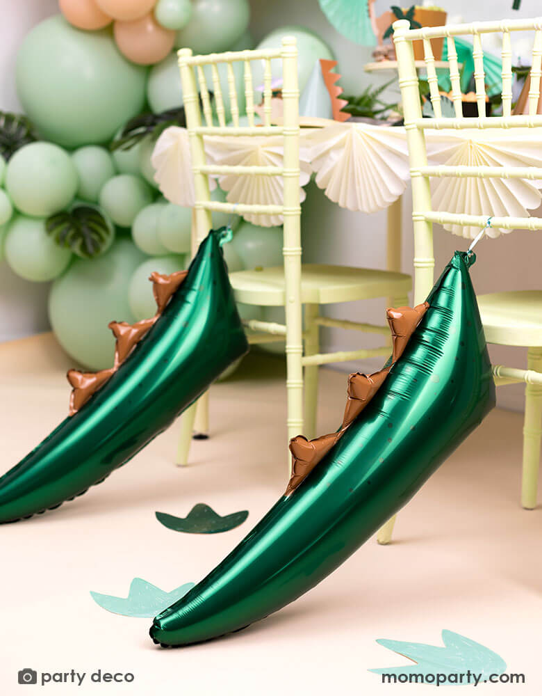 A kid's dinosaur themed birthday party table featuring party chairs decorated with Momopartys's 29" x 10" dark green dinosaur tail shaped foil balloon. On the floor there are dinosaur footprint stickers to set a prehistoric scene for a kid's dinosaur themed bash. With mint and blush balloon garland in the back this makes a perfect inspiration for a THREE-REX third birthday party bash! 