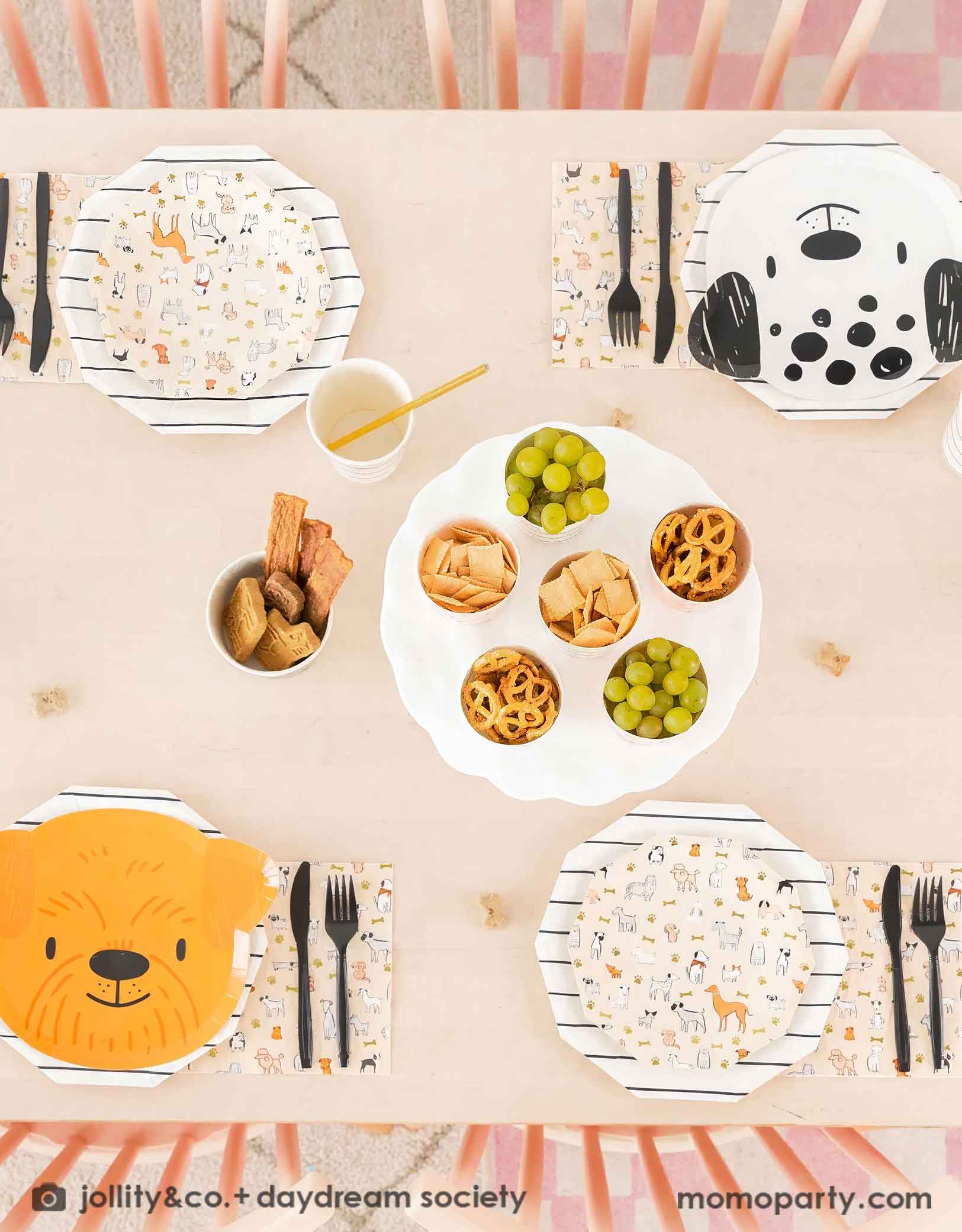 An overhead shot of Momopartys's Bow Wow Dogs themed party tablewares featuring the puppy head shaped Paper plates and Bow Wow dog small plates with black striped paper plates and cups with gold foil straw. With black utensils and some kid's snacks including grapes, pretzels, chips on the table, these warm neutral color themed modern Dog Birthday Party Supplies are perfect for a dog lover, a dog themed birthday party for kids.