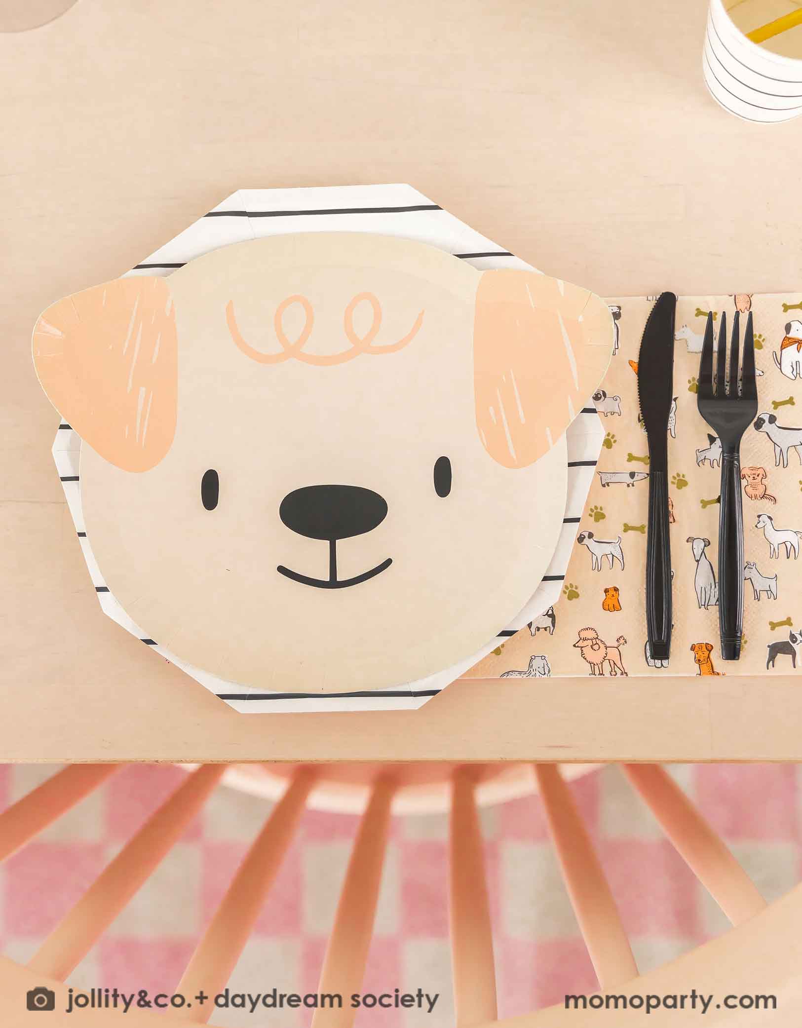 A beige dining table featuring Momopartys's cute dogs head shaped paper plate paired with a black striped paper plate, along with a napkin in modern illustrations with a warm neutral color palette of dog illustrations, with black utensil set. These modern Dog Birthday Party Supplies are perfect for a dog lover, a dog themed birthday party, Let's Pawty birthday party, adorable puppy themed party.