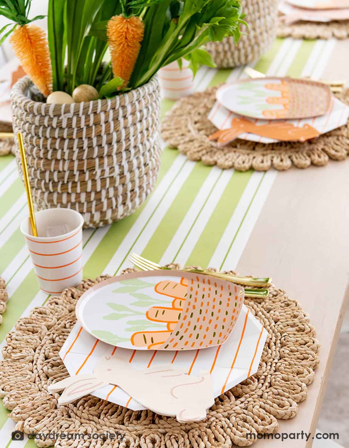 An Easter bunny themed table setting with Momopartys's bunny themed tablewares including carrot basket plates, bunny shaped napkins and matching cups and utensils on a straw-weaved placemats over a green and white striped table runner. In the center there's a green and carrot floral arrangement as the centerpiece.