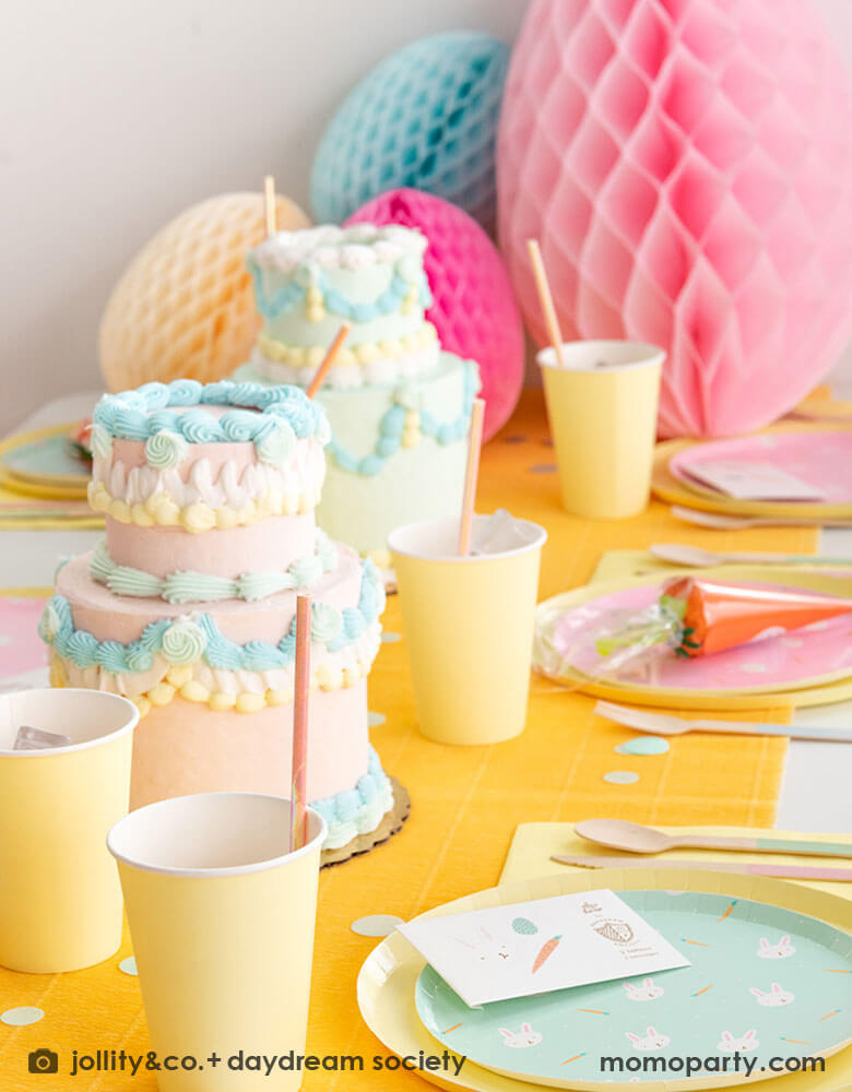 A kid's Easter party table featuring Momopartys's Easter Fun party collection by Daydream Society including Easter Fun plates in mint and pink, Easter Fun temporary tattoos, pale yellow dinner plates and party cups. On the table there are two 2-tier vintage inspired buttercream cakes with pastel piping. In the back there are multiple giant honeycomb Easter egg decorations in pastel colors of pink, ivory, and pale blue, making this a perfect inspiration for Easter brunch hosting occasion!