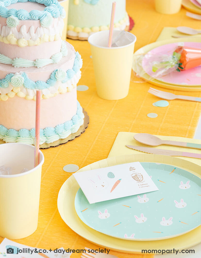 A kid's Easter party table featuring Momopartys's Easter Fun party collection by Daydream Society including Easter Fun plates in mint and pink, Easter Fun temporary tattoos, pale yellow dinner plates and party cups. On the table there are two vintage inspired buttercream cakes with pastel piping, making this a perfect inspiration for Easter brunch hosting occasion!