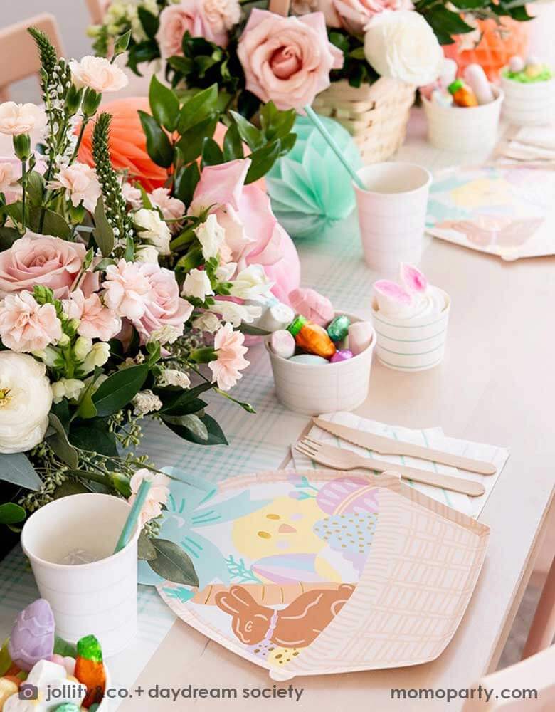 An adorable kid's friendly Easter table setting features Momopartys's Easter Fun basket shaped plates, mint striped plates, party cups by Daydream Society. In the middle there's light blue gingham table runner, Easter Egg tissue honeycomb in mint and peach and a beautiful Easter flower arrangement in a basket. On the table there's also small treat cups filled with Easter candies and chocolates, making this an adorable Easter tablescape.