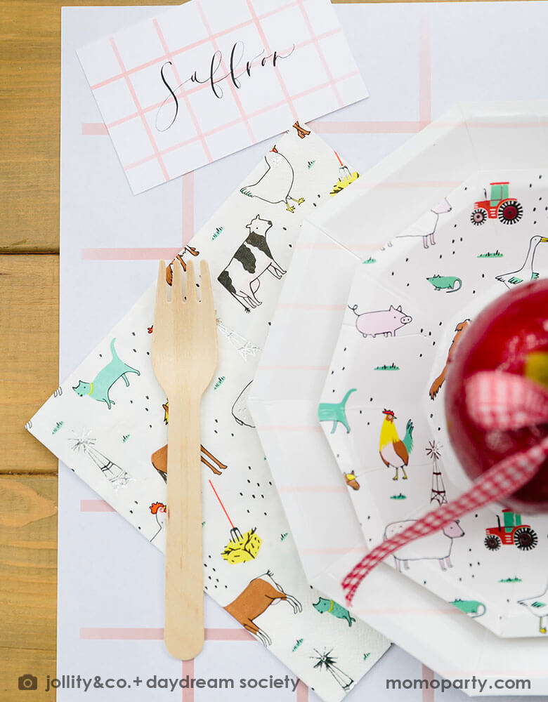 A farm themed kid's birthday party placeset featuring Momopartys's On the farm party plate & napkin paried with pink striped plates and placemat. On the plates there is a candy dipped apple adorned with red gingham ribbons. 