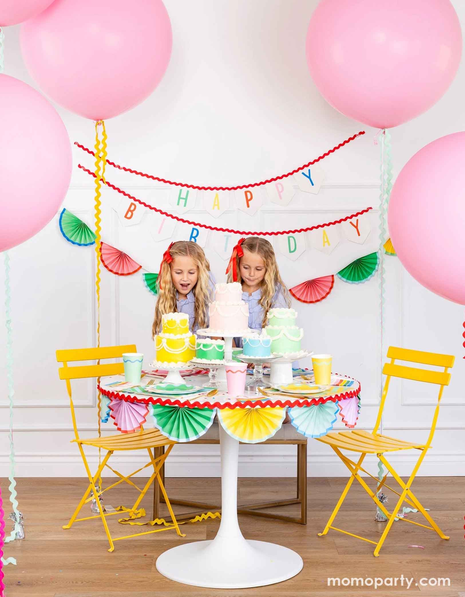 A cheerful and colorful birthday party set up featuring various festive birthday party supplies from Momopartys including rainbow colored tablewares of plates, cups, napkins on the table along with multiple pastel colored birthday cakes. The table is decorated with colorful bunting garland around and in the back of the wall hung a Happy Birthday felt garland and the colorful bunting banner. On the sides are some jumbo pink balloons floating above and two girls standing excitedly next to the cakes.