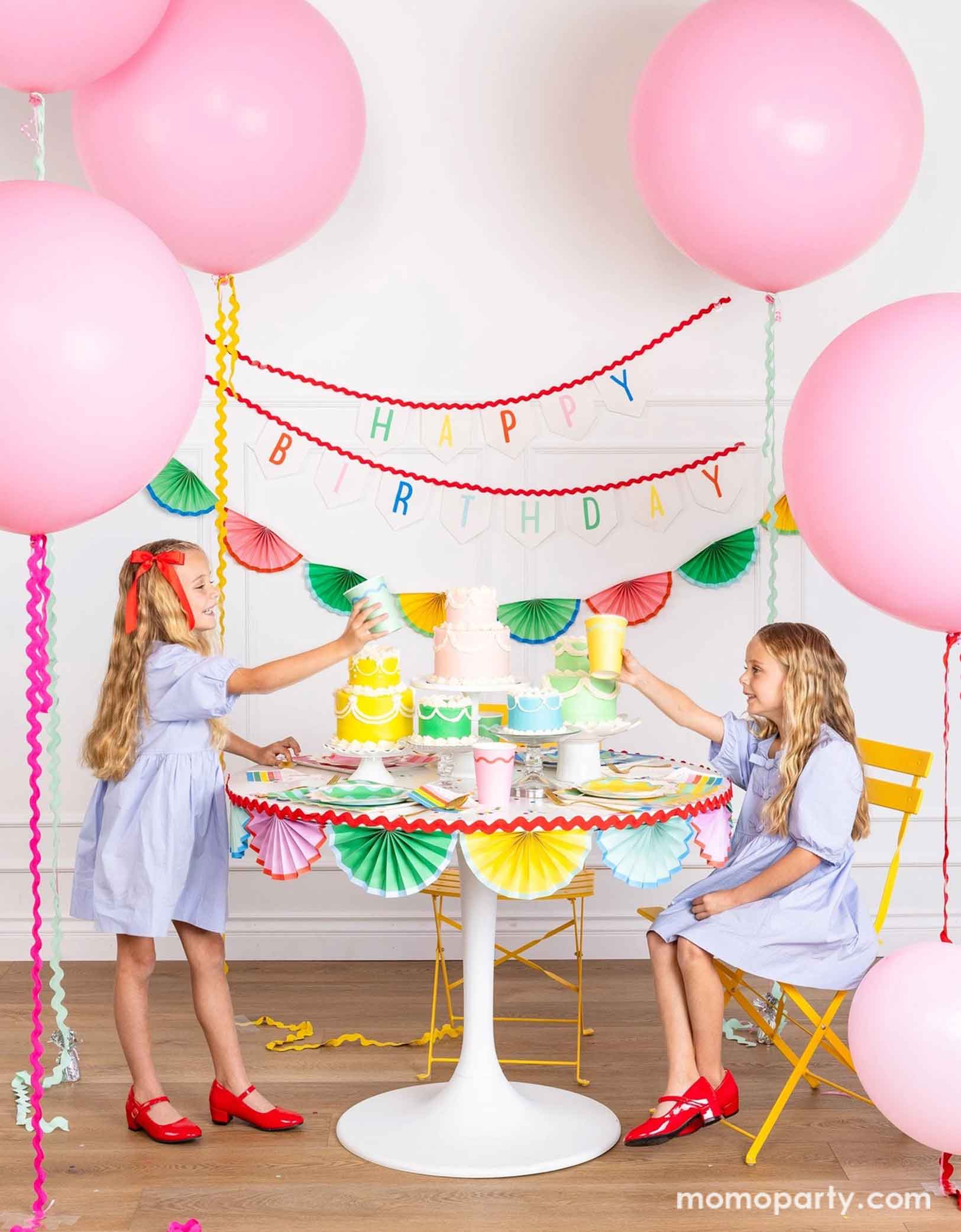 A cheerful and colorful birthday party set up featuring various festive birthday party supplies from Momopartys including rainbow colored tablewares of plates, cups, napkins on the table along with multiple pastel colored birthday cakes. The table is decorated with colorful bunting garland around and in the back of the wall hung a Happy Birthday felt garland and the colorful bunting banner. On the sides are some jumbo pink balloons floating above and two girls  sitting at the table and holding cups.