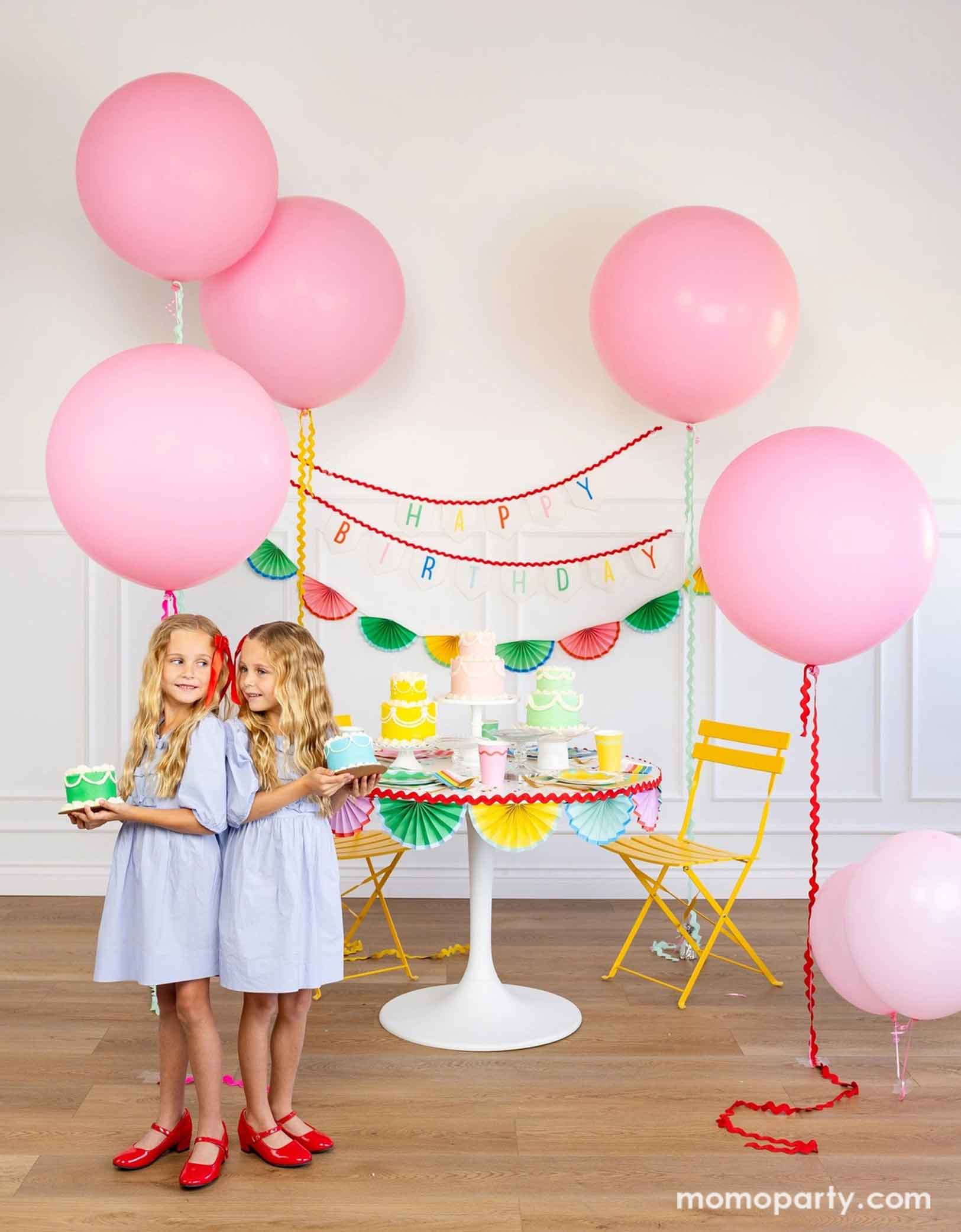 A cheerful and colorful birthday party set up featuring various festive birthday party supplies from Momopartys including rainbow colored tablewares of plates, cups, napkins on the table along with multiple pastel colored birthday cakes. The table is decorated with colorful bunting garland around and in the back of the wall hung a Happy Birthday felt garland and the colorful bunting banner. On the sides are some jumbo pink balloons floating above and two girls standing in the front holding birthday cakes.