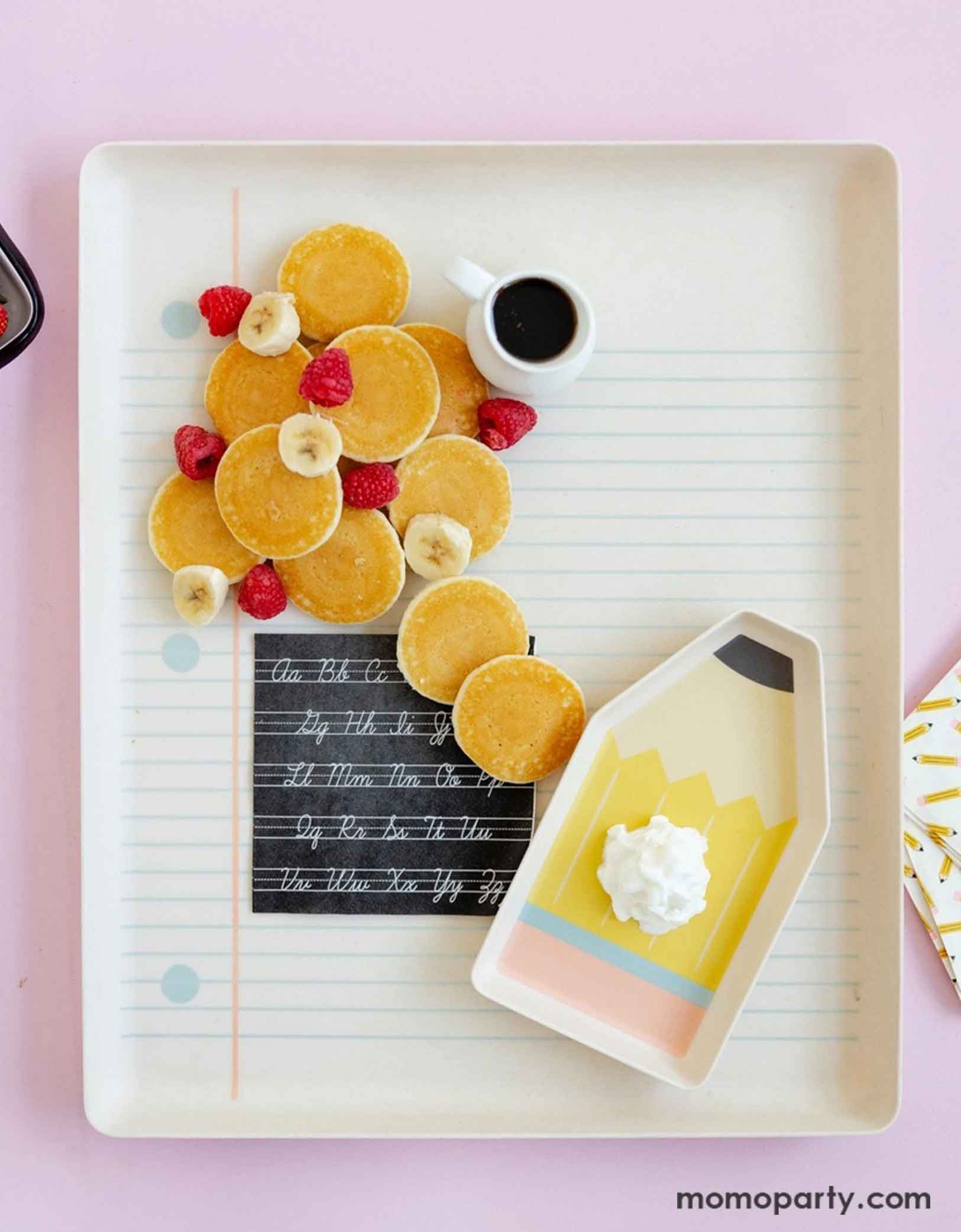 An overhead shot of Momopartys's 17" x 14" Paper and Pencil Bamboo Tray Set filled with a bunch of petite pancakes, banana bites, raspberries and a small syrup pourer. On the notebook bamboo tray is the black Cursive Abcs Paper Small Napkin and the pencil shaped bamboo try with a dollop of whip cream. This makes a great inspiration idea for a special first day of school breakfast for kids to celebrate the new school year!