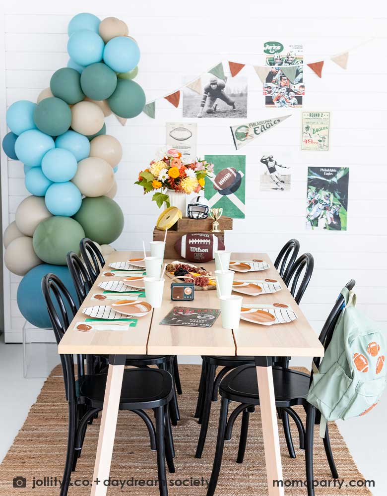 A kid's football birthday party set up with a modern party table filled with football themed tableware from Momopartys including the die-cut football shaped plates, black striped plates, green and mint napkins and party cups. In the center of the table there's a cheese charcuterie board and some decorations like a football, a trophy and a football helmet as the centerpiece. On the wall there are some old school football party banners, posters and party balloons to add some festivities to the celebration!