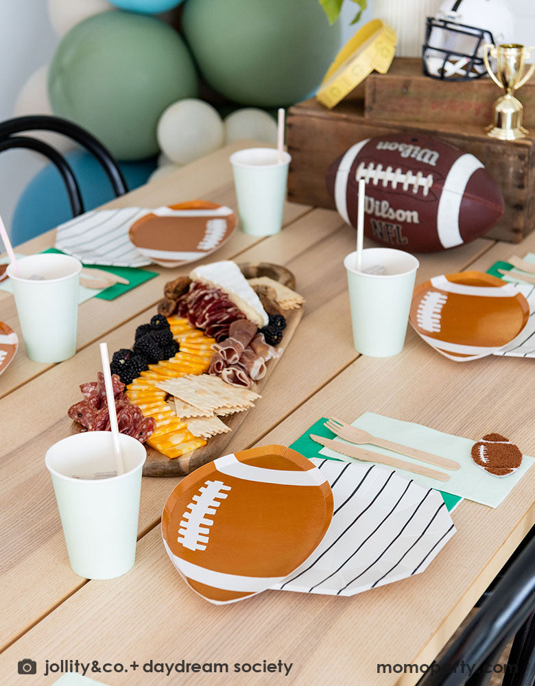 A modern kid's football party table filled with Momopartys's football themed party tableware by Daydream Society including the die-cut football shaped plates, black striped plates, green and mint napkins and party cups. In the center of the table there's a cheese charcuterie board and some decorations like a football, a trophy and a football helmet as the centerpiece - a great inspirations for football themed celebrations! 
