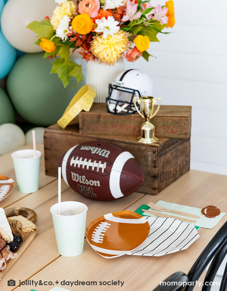 A modern kid's football birthday party table filled with Momopartys's football themed party tableware by Daydream Society including the die-cut football shaped plates, black striped plates, green and mint napkins and party cups. In the center of the table there's a cheese charcuterie board and some decorations like a football, a trophy and a football helmet as the centerpiece. On the wall there are some old school football posters and color matching balloons to add some festivities to the celebration!
