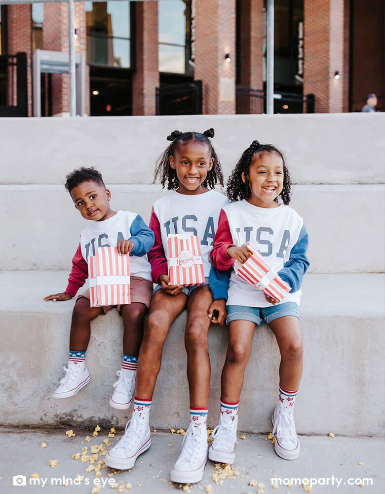 Three children dressed their USA sweatshirt sitting on a bench eating Cracker Jacks of Momopartys's Stars and Stripes Treat Bags by My Mind's Eye featuring red striped design with "Hip Hip Hooray" on it, it's perfect for at this year's Fourth of July parade viewing!