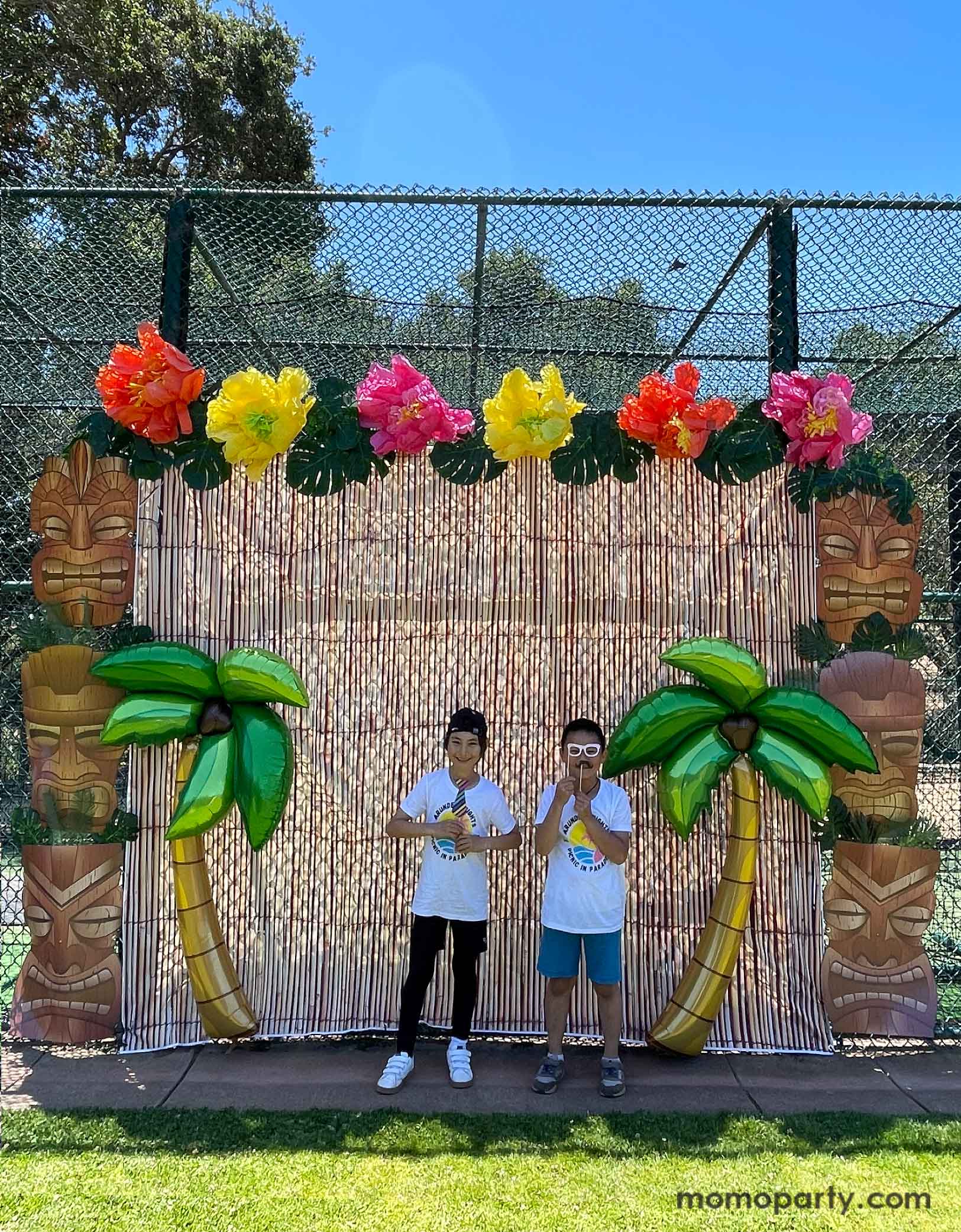 Two school aged boys in a  luau,, tropical, tiki themed party photo backdrop decoration set up by Momopartys featuring bamboo party backdrop, jumbo Tiki Mask Cardboard Cutouts adorned with palm leaves as two pillars. On the top of the backdrop are some tissue paper Hawaiian hibiscus flowers in orange, yellow and pink colors along with palm tree leaf decorations. In the front are two 65" Jumbo Palm Tree Shaped Foil Balloons, making this a festive decoration idea for kid's island themed birthday celebration.