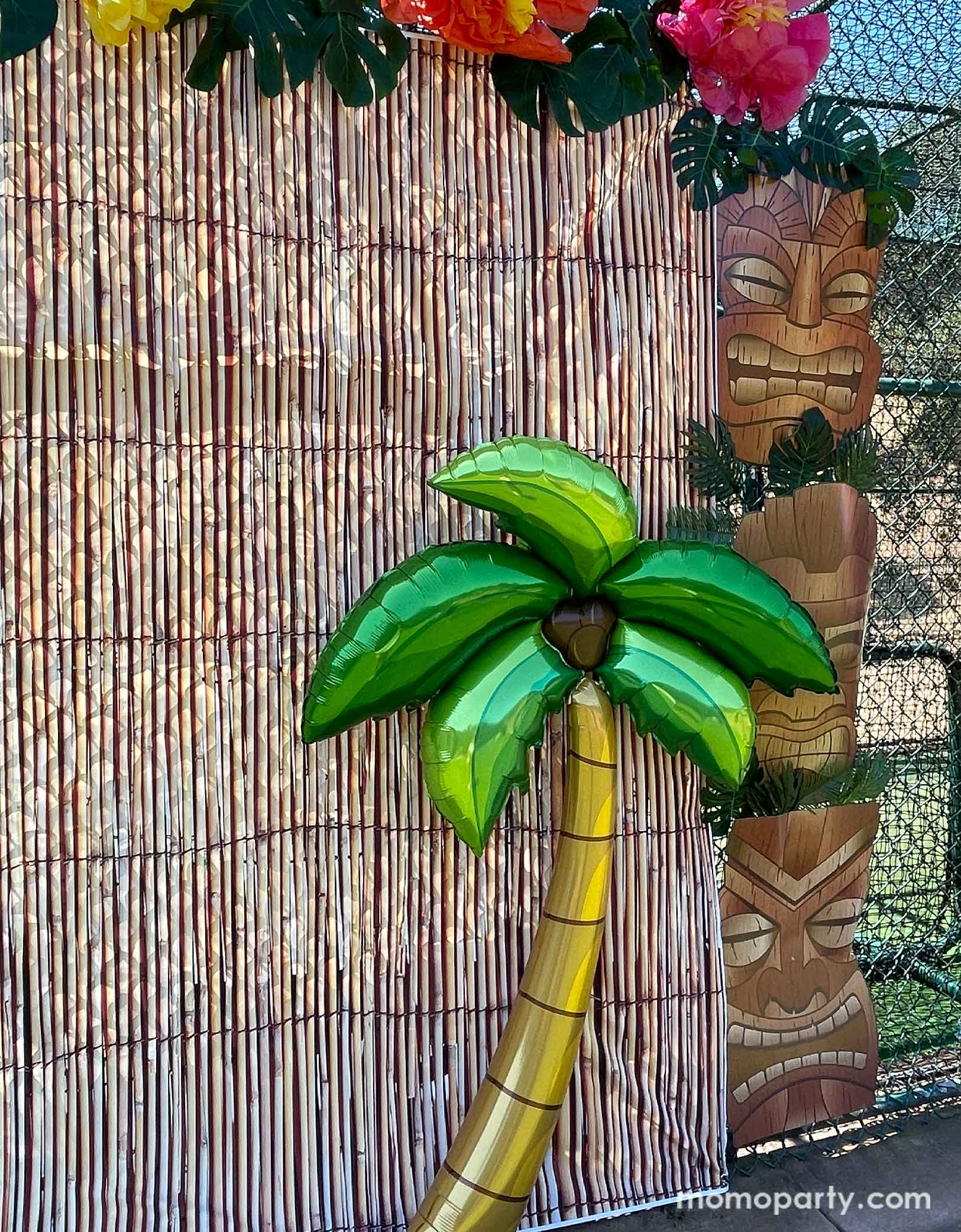 Momo Party's 65" Jumbo Palm Tree Shaped Foil Balloon in  in a luau, tropical, tiki themed party photo backdrop decoration featuring bamboo party backdrop, jumbo Tiki Mask Cardboard Cutouts adorned with palm leaves as two pillars. On the top of the backdrop are some tissue paper Hawaiian hibiscus flowers with palm tree leaf decorations. This palm tree shaped foil balloon is an easy and fun decoration to set a island scene for any tropical themed celebration.