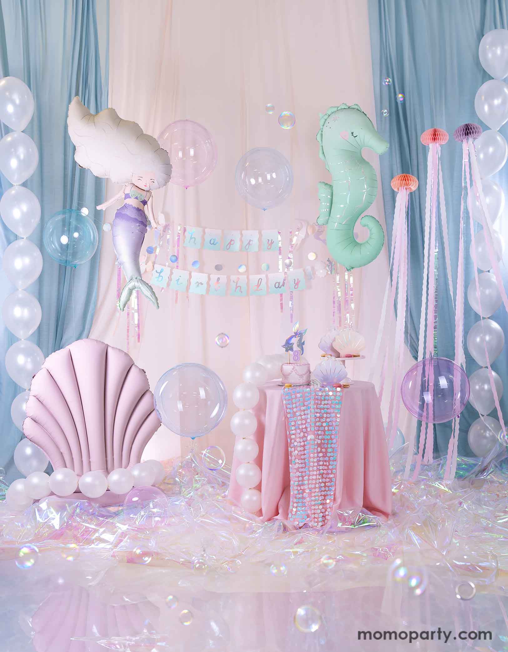 An enchanting and dreamy kid's mermaid party set up featuring Momopartys's under the sea mermaid themed party supplies including a floating mint seahorse balloon, a mermaid shaped balloon, a lilac seashell standing foil balloon adorn with pearl white balloons, the mermaid happy birthday part garland, and a table with mermaid themed tableware. With a white and light blue curtain in the back as the backdrop, lots of see-through bubble balloons and an iridescent cellophane on the floor.