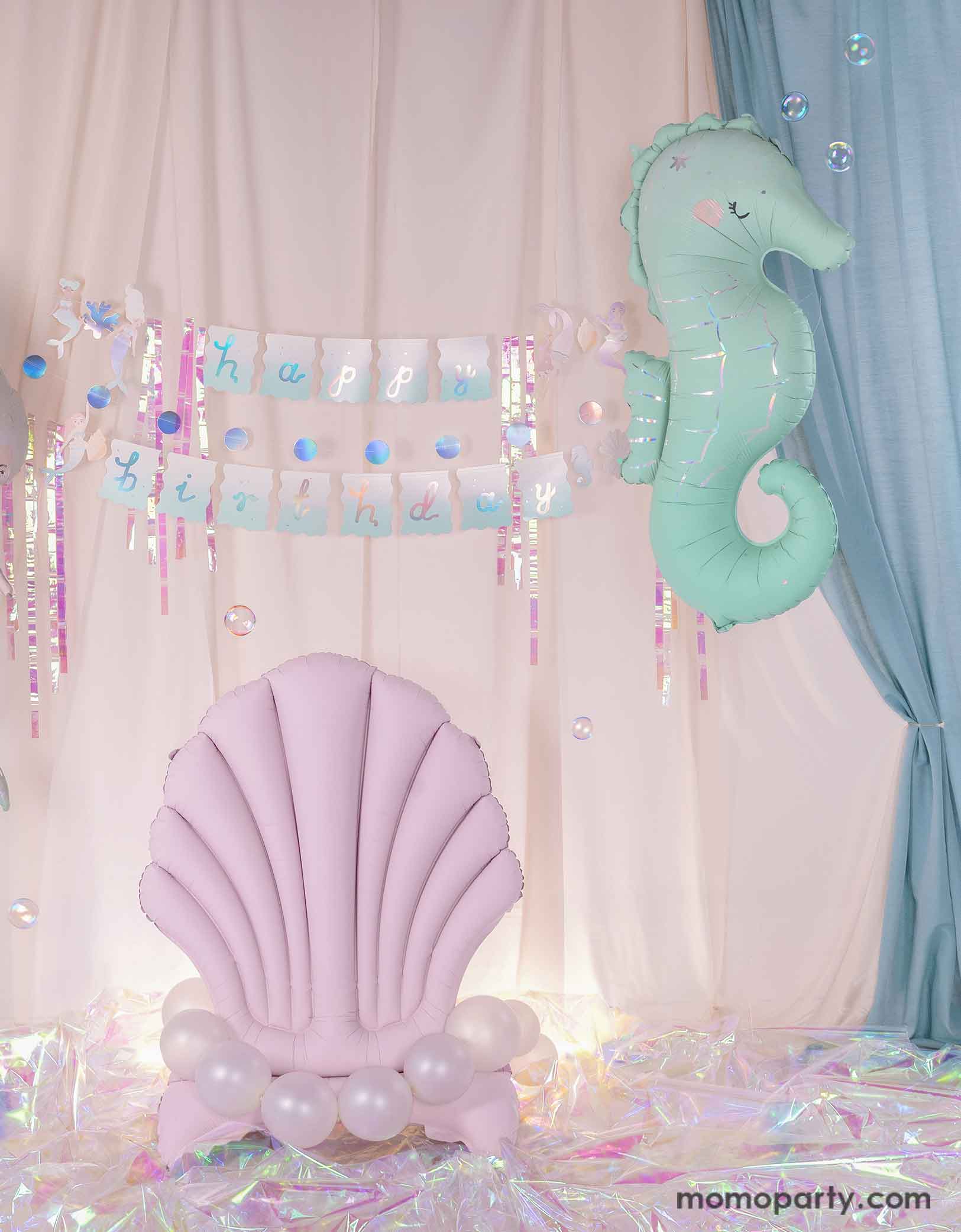 An enchanting and dreamy kid's mermaid party decoration set up featuring Momopartys's under the sea mermaid themed party supplies including a floating mint seahorse balloon, the lilac seashell standing foil balloon adorn with pearl white balloons and the mermaid happy birthday part garland set with iridescent streamers. With a white and light blue curtain in the back as the backdrop and iridescent cellophane on the floor, making this a magical setting for kid's mermaid party.