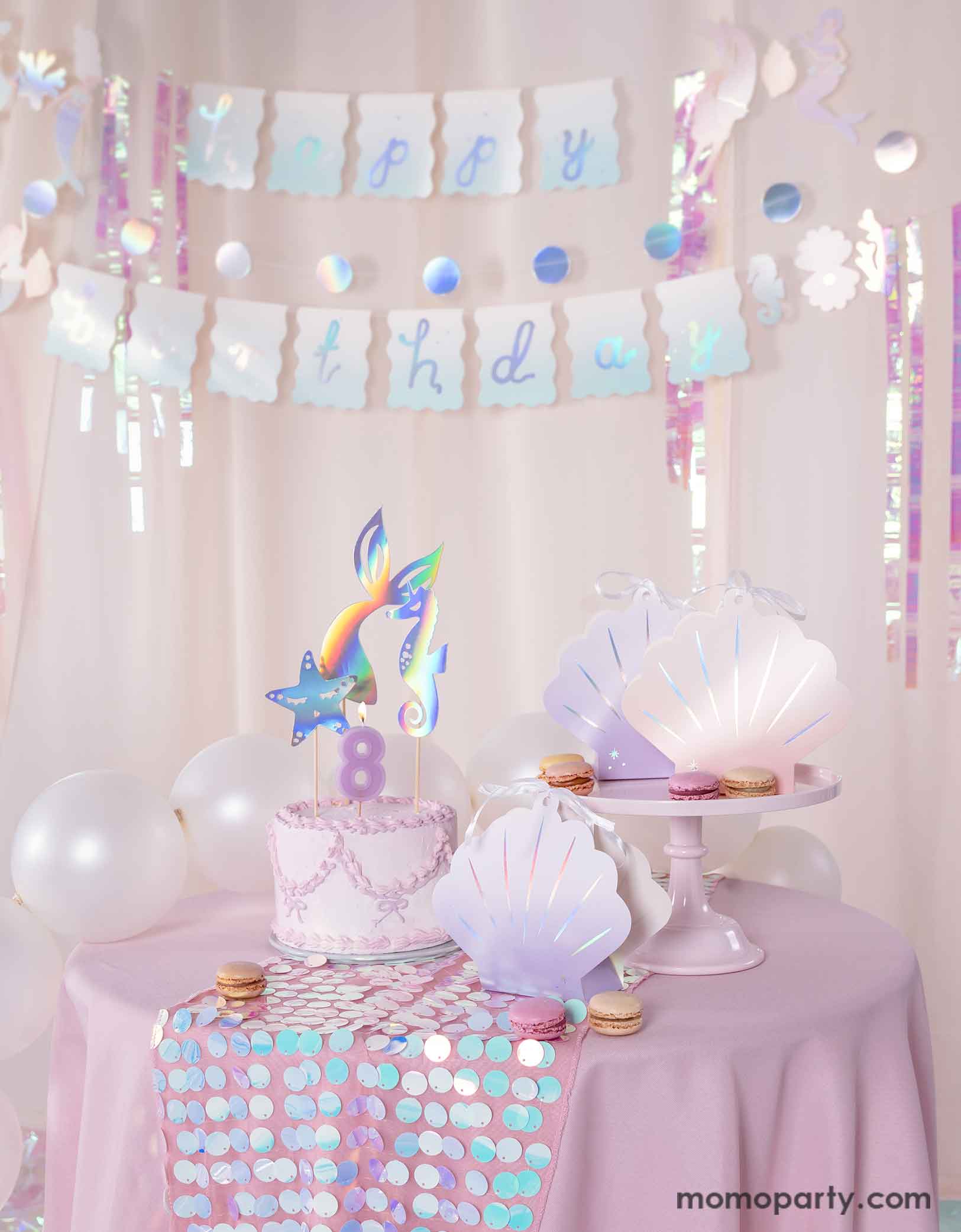 A mermaid birthday party table set uo featuring various mermaid themed party supplies from Momopartys including the  ombre pink and lilac shell shaped treat boxes, a pink butter cream cake topped with mermaid themed toppers. Behind the table there's a pink curtain adorned with the mermaid happy birthday party banner as the backdrop. Along with pearl white balloons around, creating a dreamy and whimsical scene for a magical under the sea bash. 