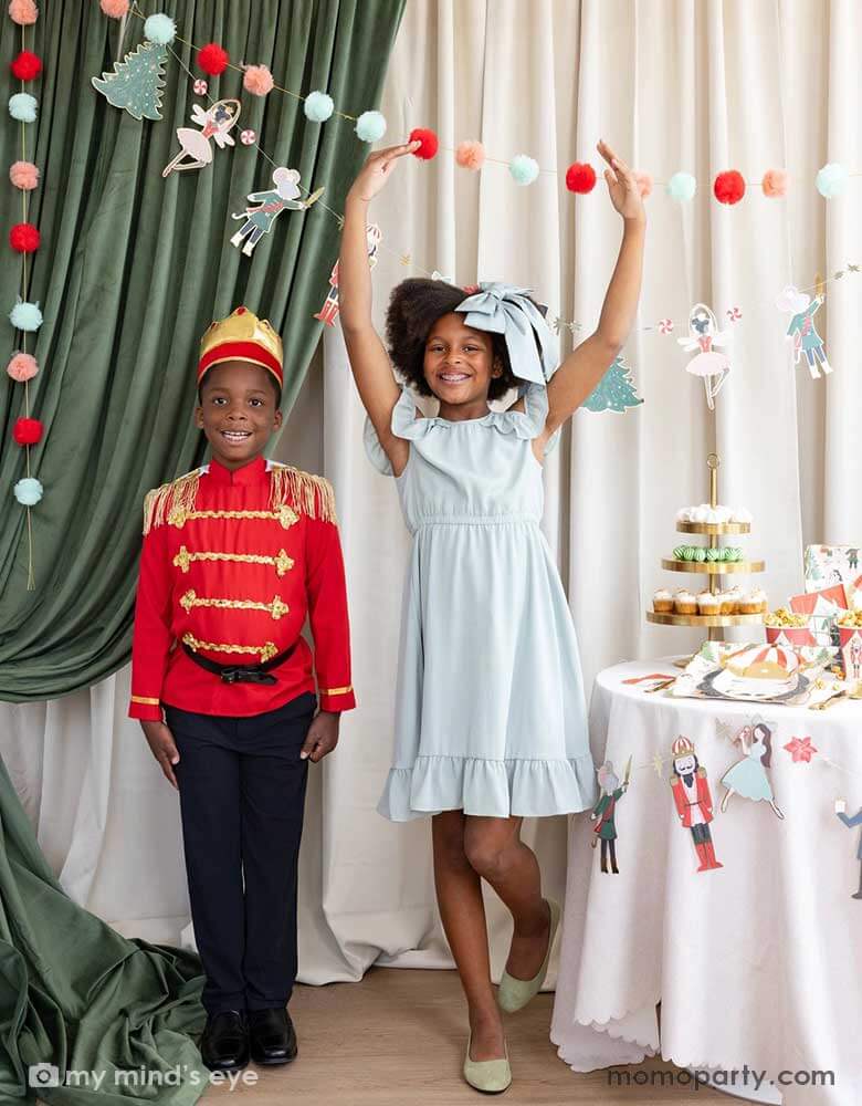 A girl and a boy dressed as the Nutcracker characters in a Nutcracker themed holiday party. The girl is doing a ballet dance pose and they're standing next to a party table filled with Holiday treats and Nutcracker themed party supplies. In their back there's a stage curtain in satin green and beige which is decorated with Momopartys's Nutcracker 6' garland with pom-poms which adds a festive touch to the Holiday decoration.