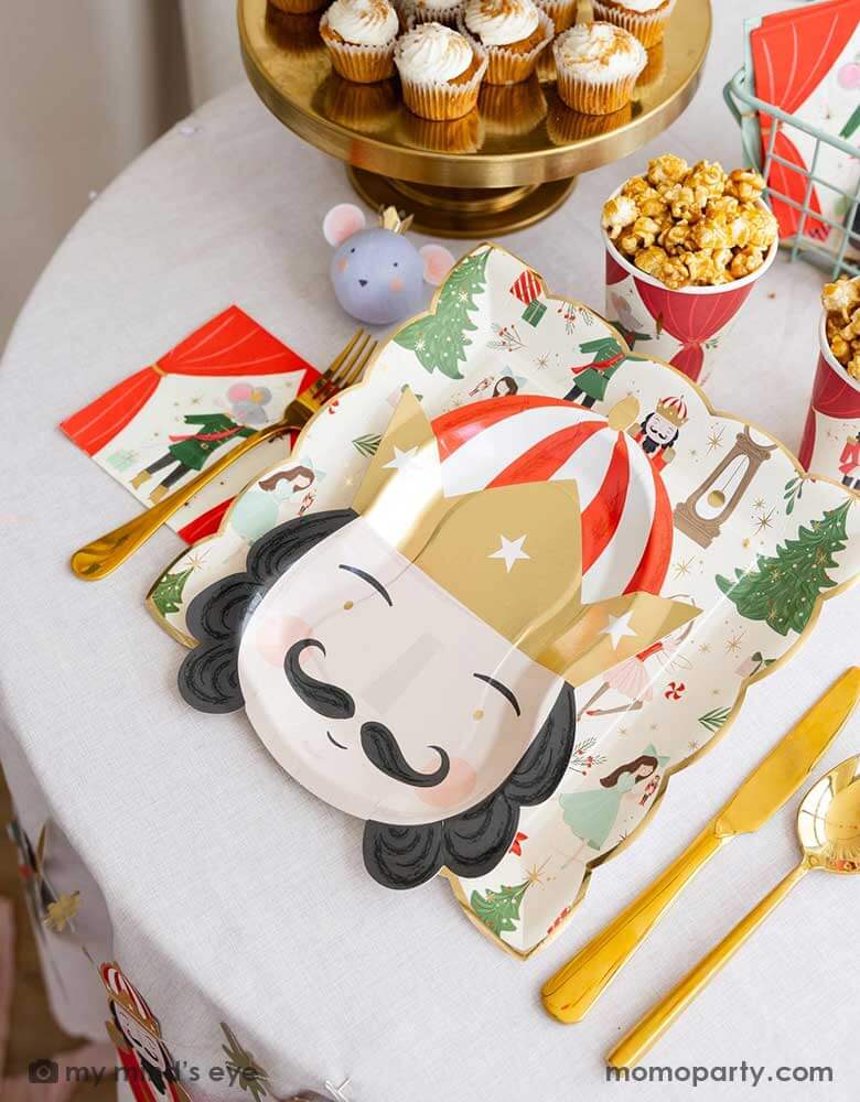 A festive Holiday party table featuring Momopartys's Nutcracker themed tableware including the Nutcracker pattern plate, Nutcracker head shaped plate, Nutcracker small napkins, Nutcracker party cups and the Nutcracker mouse surprise ball by My Mind's Eye. On the party table there are also some kid's friendly treats including cupcakes and popcorns - making this a perfect inspo for kid's Nutcracker themed Holiday celebration this Christmas.