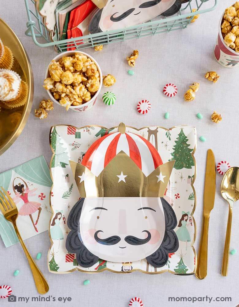 A festive Nutcracker themed Holiday party tablescape featuring Momopartys's Nutcracker party supplies including the Nutcracker pattern plate, Nutcracker head shaped plate, Nutcracker napkins, party cups which are filled with popcorn. On the table there are some Holiday treats including  green and red candies, peppermint and cupcakes making this a festive Christmas party table for kids and guests of all ages!