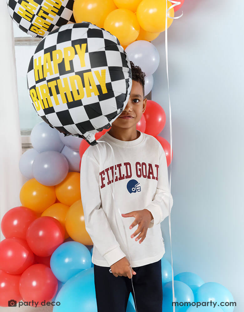 A boy holding Momopartys's 14" checkered flag foil balloon with gold foil Happy Birthday written on it. He is standing in front of a festive balloon garland in race car inspired colors, ready for a fun race car birthday bash to begin!