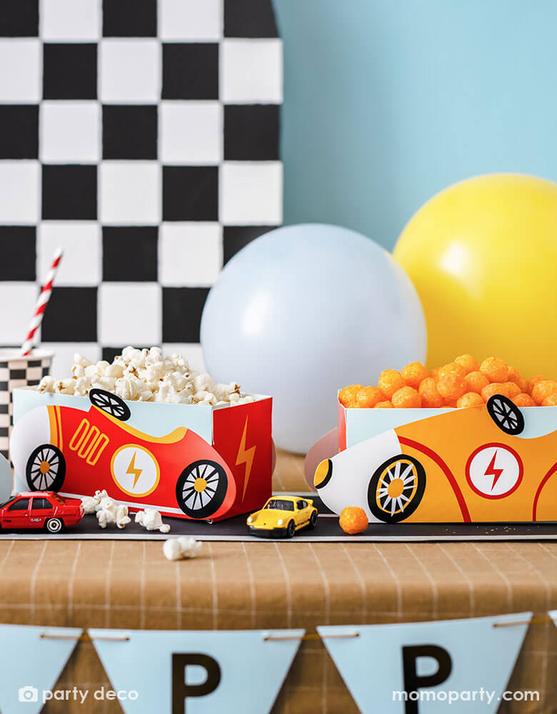 A modern kid's race car themed party table features Momopartys's race car snack boxes in yellow and red, filled with popcorn and cheese balls. On the table there are checkered pattern party cups and race track table runner, with toy cars decorated. In the back there's a checkered backdrop decorated with some party balloons in light blue and yellow, making this a perfect inspo for kid's race car themed parties including a "Two Fast" second birthday party or a "Fast One" first birthday party."
