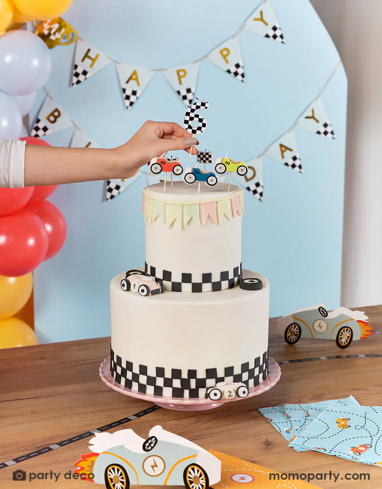 A race car themed birthday cake decorated with checkered pattern at the bottom topped with Momopartys's race car candles, a hand putting a checkered number 6 candle on the cake. In the back there's a race car themed trophy happy birthday banner hung on the wall. On the table there are some race car themed paper napkins and race car shaped party invitation. Makes it a great inspiration for kid's race car birthday party.