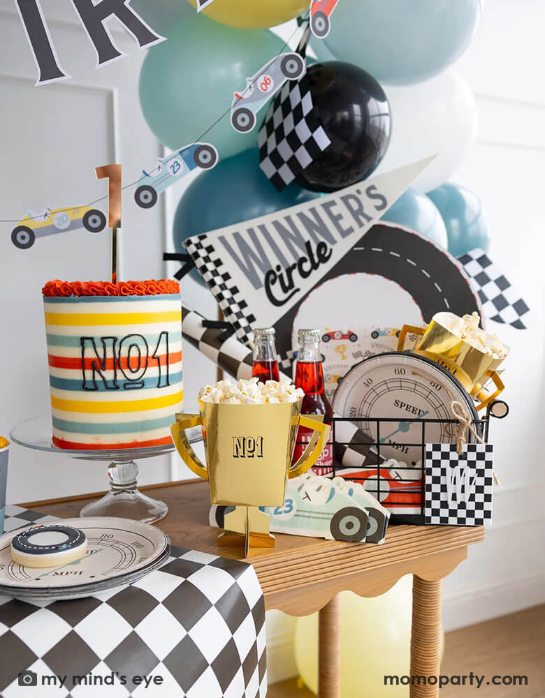 A party bin filled with race car themed party supplies from Momopartys including race track placemats, speedometer shaped paper plates, checkered table runner, race car pattern square plates, race car shaped napkins, and a felt "Winner's Circle" party pennant. Around the basket is a colorful buttercream cake and some party balloons decorated behind the party table, with a gold trophy treat box with popcorn along with some cake plates. Making this a perfect inspo for kid's race car themed celebrations.