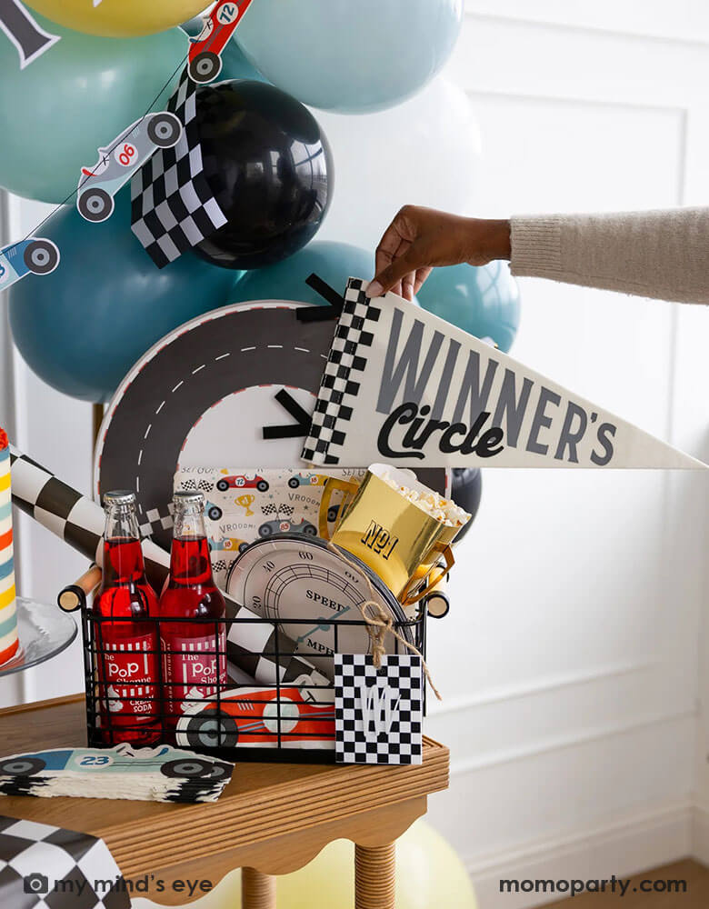 An occasion bin filled with race car themed party supplies from Momopartys including race track placemats, speedometer shaped paper plates, checkered table runner, race car pattern square plates, race car shaped napkins, and trophy shaped treat boxes. A hand is reaching to grab the felt "Winner's Circle" party pennant from the basket. Around the basket is a colorful buttercream cake and some party balloons and checkered flags decorated behind the table - a perfect inspo for kid's race car themed parties.