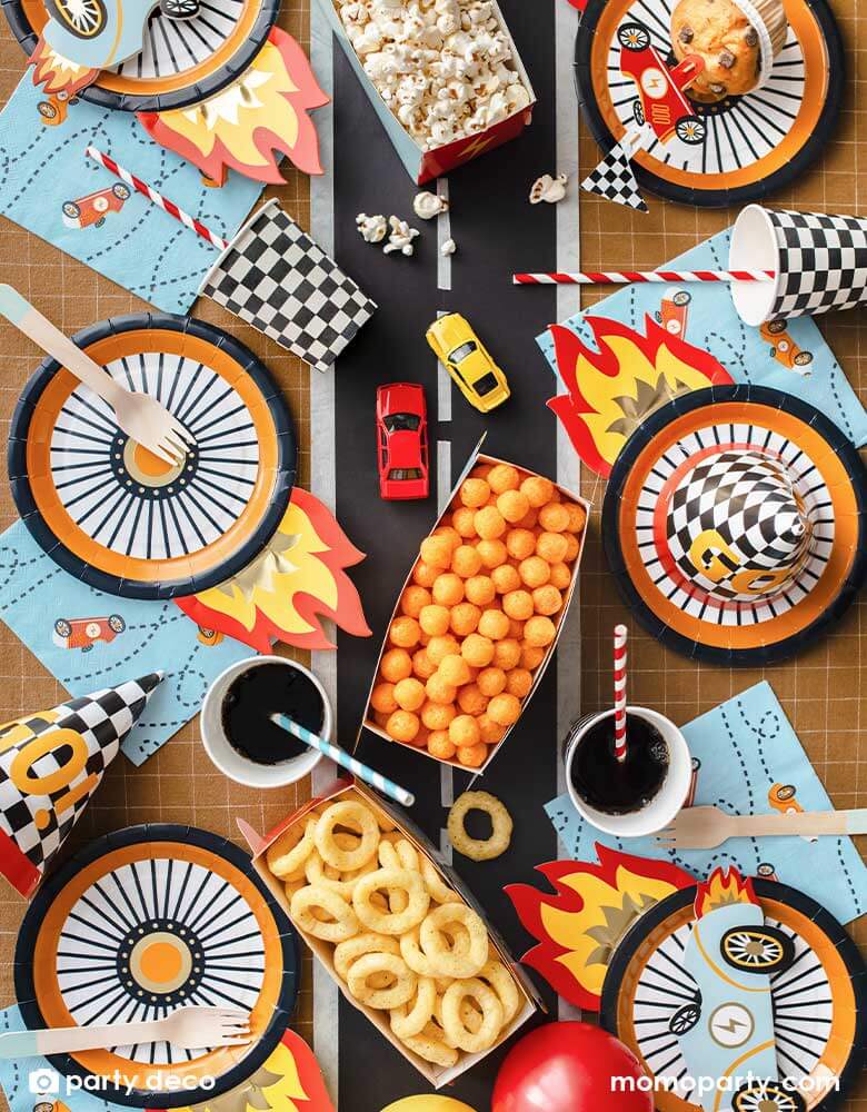 A kid's race car themed party table filled with race car themed party supplies from Momopartys including the wheel shaped plates, pastel blue race car large napkin, and checkered party cups. On the table there are muffins topped with race car themed cupcake toppers and a red vintage race car shaped snack holder filled with popcorn. All laid on a brown checkered tablecloth adorned with race car track in the middle with some party snacks and toy race cars, making these a perfect inspo for a car themed party!