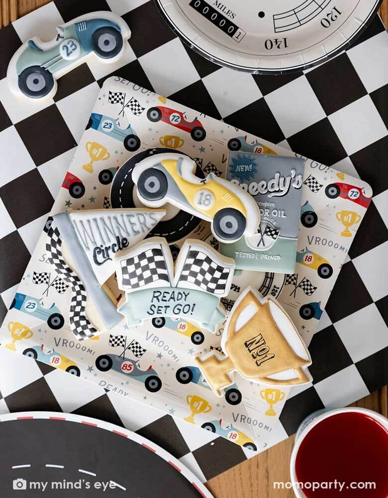 A close-up shot of a race car party table featuring Momopartys's race car pattern square paper plates, speedometer shaped plates and checkered table runner. On the race car pattern plate, there are multiple race car themed sugar cookies with matching style and colors, making this a perfect inspo for kid's race car themed birthday celebration, be it a "Two Fast" second birthday party, a "Fast One" first birthday party.