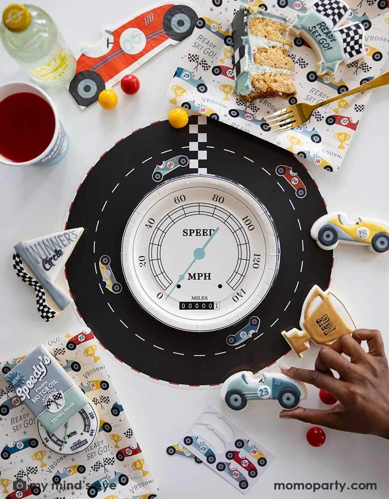 A fun race car themed party table place set featuring adorable race car themed party supplies from Momopartys including a race track placemats adorned with interactive race car stickers in the middle of the placemat is a speedometer shaped plate. Around the tableset laid some race car themed sugar cookies in matching colors and designs around the race car pattern square plates by My Mind's Eye. A perfect inspo for kid's race car themed party tablesetting ideas.