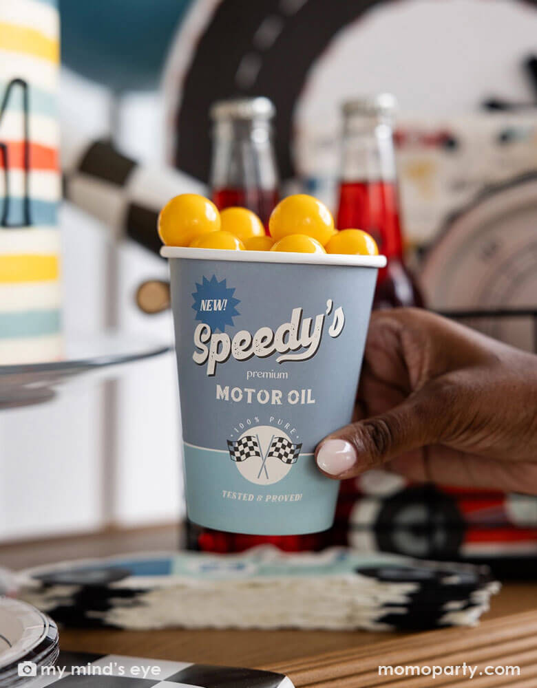 A kid's race car party featuring Momopartys's 12 oz race car cup by My Mind's Eye. Comes in a set of 8 paper cups in light blue and mint with Speedy's motor oil logo on it and some checkered flags design. In the car there are yellow gum balls as kid's treats and in the back you can see some festive race car themed party decorations, birthday cakes, treats and drinks.