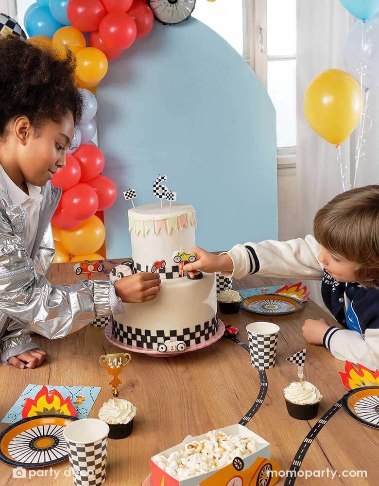Two kids putting race car shaped candles on a race car themed birthday cake, around the cake there are lots of race car themed party supplies including Momopartys's wheel shaped plates, napkins, checkered party cups, cupcakes topped with race car themed toppers, and race car shaped snack holders. In the back there's a pastel blue backdrop adorned with colorful balloon garland in race car themed colors.