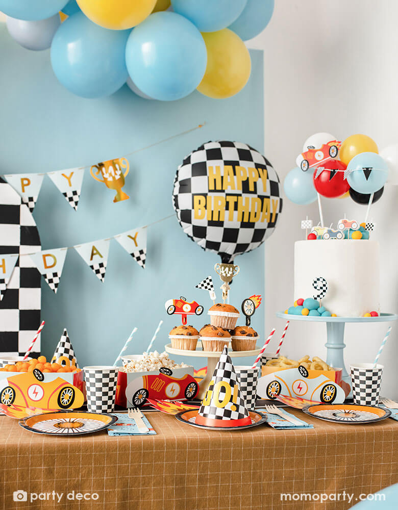 A kid's race car themed party table filled with race car themed party supplies from Momopartys including wheel shaped plates, checkered flag GO! party hats, blue race car large napkin, and checkered party cups. On the table there are muffins topped with race car themed cupcake toppers and vintage race car shaped snack holder filled with popcorn and onion ring chips. Above the table, there's a race car flag themed birthday banner and checkered happy birthday foil balloon.