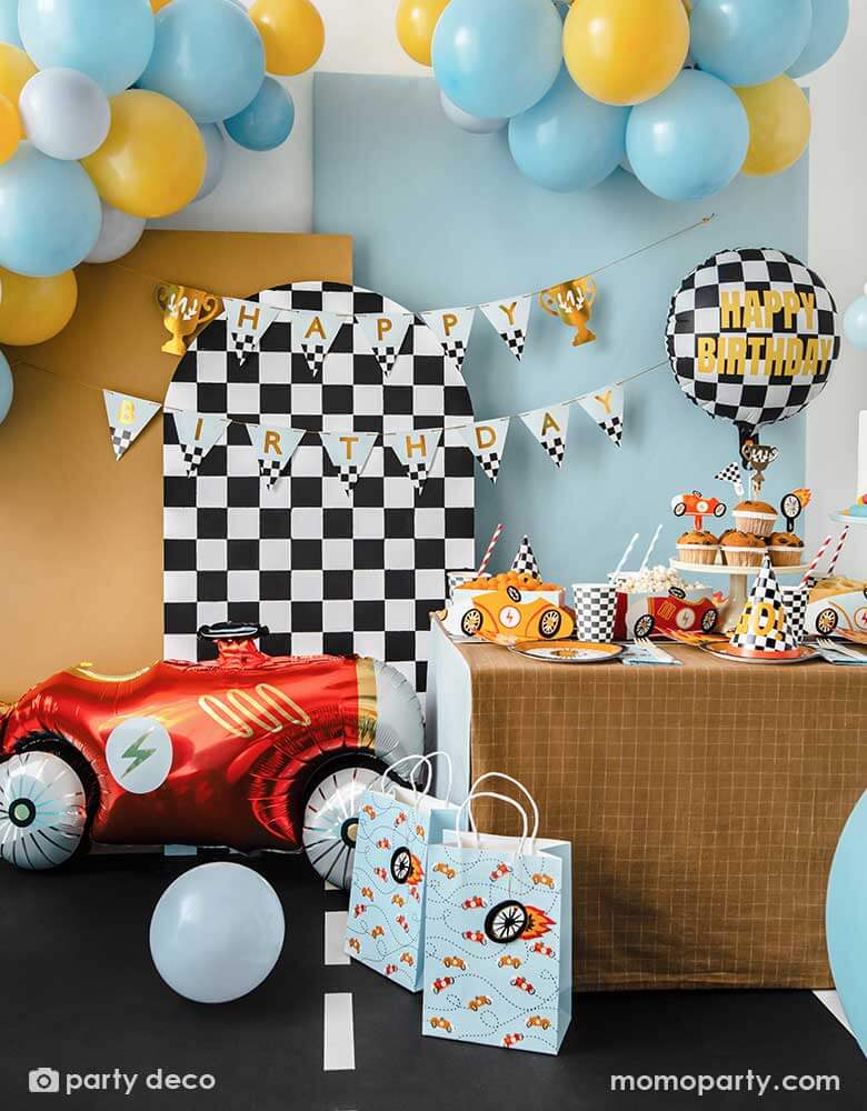 A kid's race car themed party featuring a table filled with race car themed party supplies from Momopartys including wheel shaped plates, checkered flag GO! party hats, blue race car large napkin, and checkered party cups. Behind the table is a checkered party backdrop adorned with trophy happy birthday banner with colorful balloon garland. On the floor there's a vintage red race car foil balloon setting the scene for this festive celebration. A perfect inspo for kid's Two Fast second birthday party.