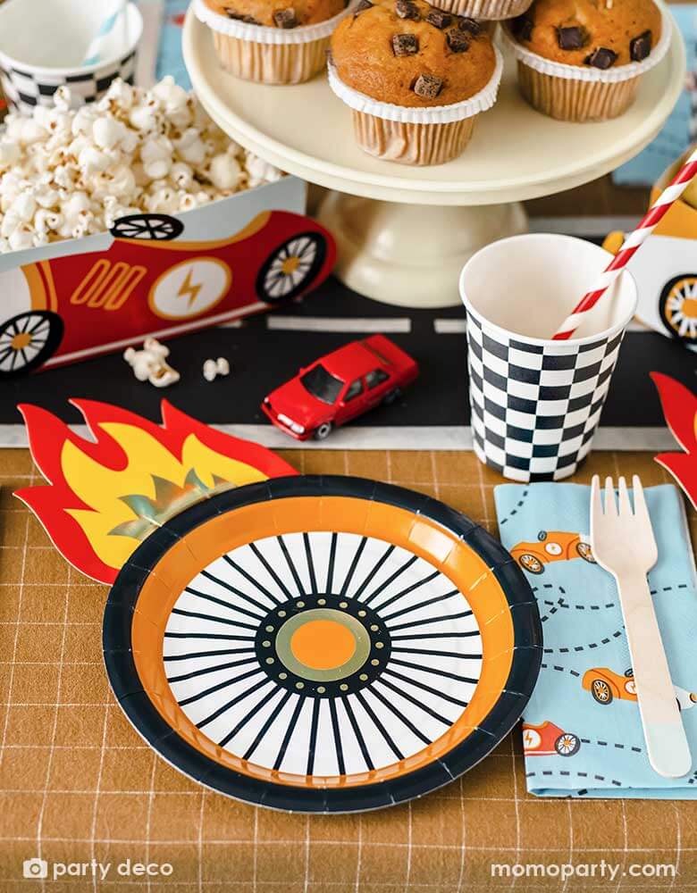 A kid's race car themed party table filled with race car themed party supplies from Momopartys including wheel shaped plates, pastel blue race car large napkin, and checkered party cups. On the table there are muffins topped with race car themed cupcake toppers and a red vintage race car shaped snack holder filled with popcorn. All laid on a brown checkered tablecloth adorn with race car track in the middle with some toy race cars, making these a perfect inspo for a rad kid's race car birthday party!