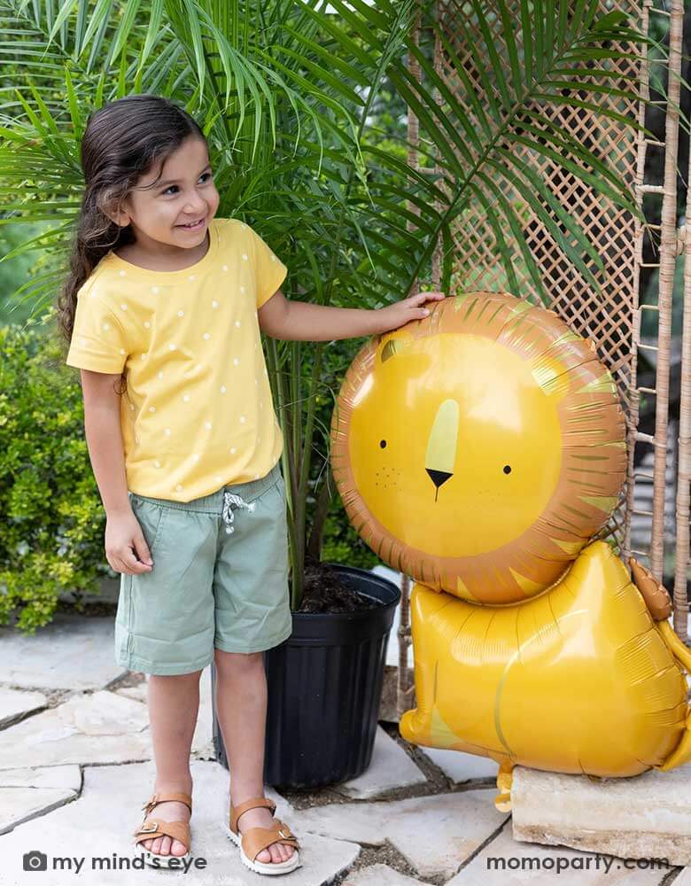 A little girl dressed in her safari adventure themed outfit standing next to Momopartys's 24" lion shaped foil mylar balloon. This charming lion shaped balloon adds the perfect amount of wild to a birthday celebration that will have your guests exclaiming "Oh My!"