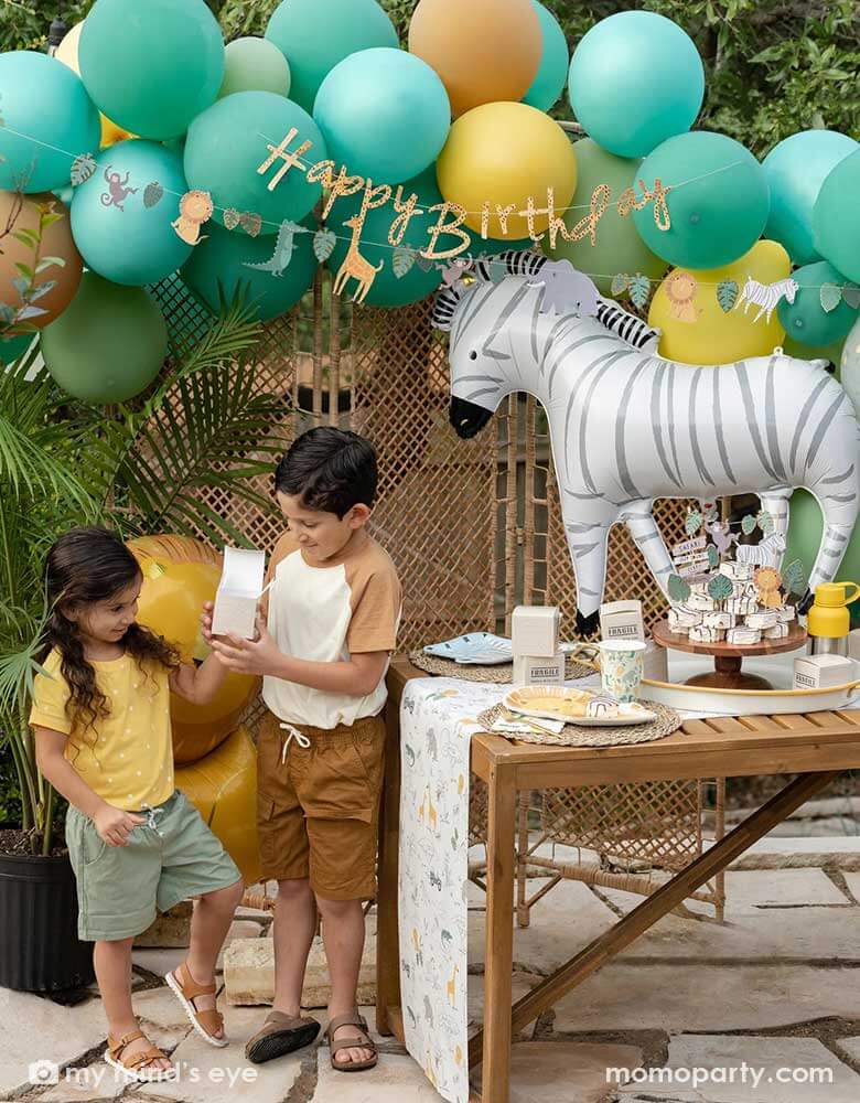 A festive kid's safari themed birthday party set up feature Momopartys's 24" zebra shaped foil mylar balloon by My Mind's Eye. Above it is a safari themed balloon garland in green, orange and yellow adorned with safari animal happy birthday banner. Next to the table which features multiple safari themed tableware and some zebra cakes in the middle are two kids in safari themed outfits opening Momopartys's cargo party favor boxes for some goodies.