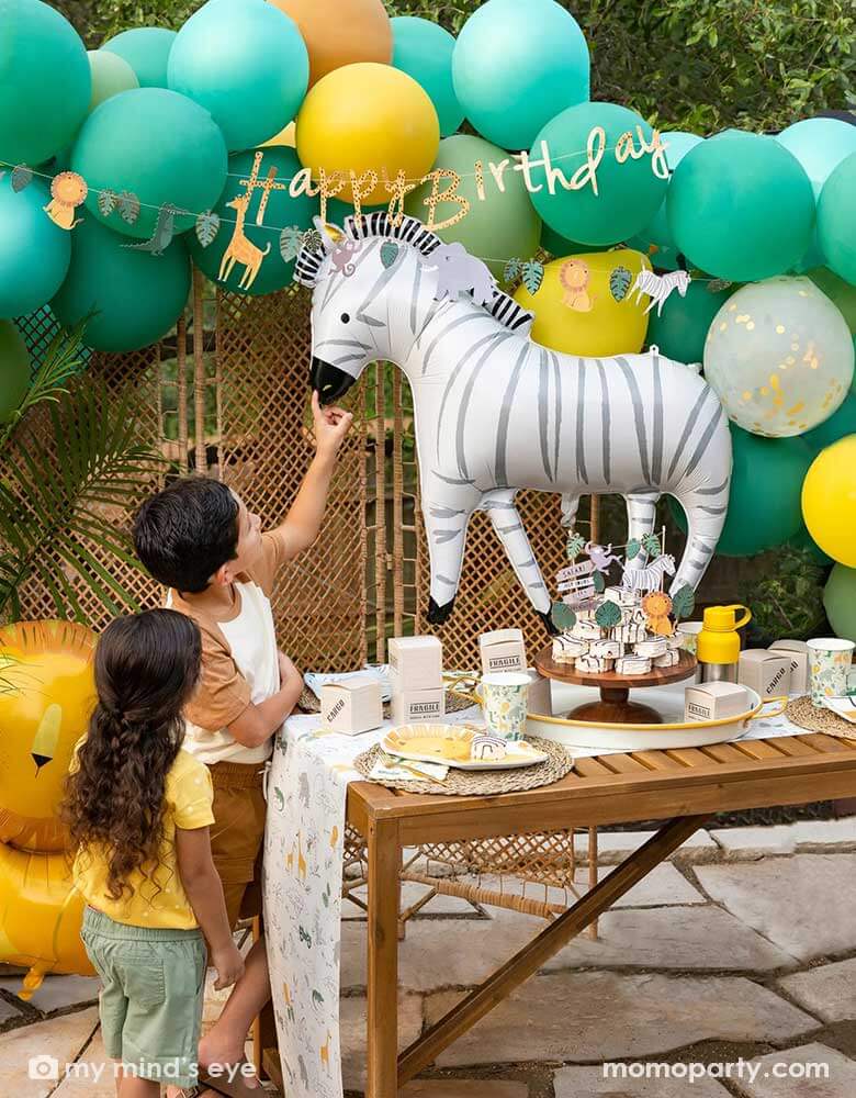 A festive kid's safari themed birthday party set up feature Momopartys's 24" zebra shaped foil mylar balloon by My Mind's Eye. Above it is a safari themed balloon garland in green, orange and yellow adorned with safari animal happy birthday banner. Next to the table which features multiple safari themed tableware and some zebra cakes in the middle are two kids pretending to feel the zebra and pet him.