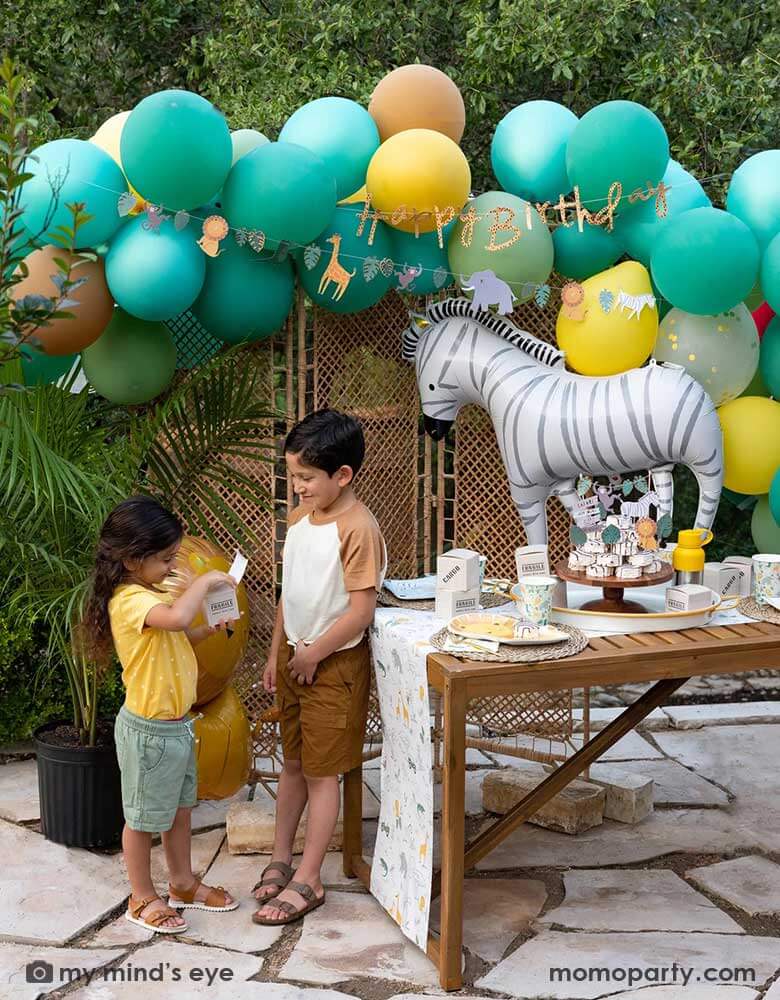 A festive kid's safari themed birthday party set up feature Momopartys's 24" zebra shaped foil mylar balloon by My Mind's Eye. Above it is a safari themed balloon garland in green, orange and yellow adorned with safari animal happy birthday banner. Next to the table which features multiple safari themed tableware and some zebra cakes in the middle are two kids in safari themed outfits opening Momopartys's cargo party favor boxes for some goodies.