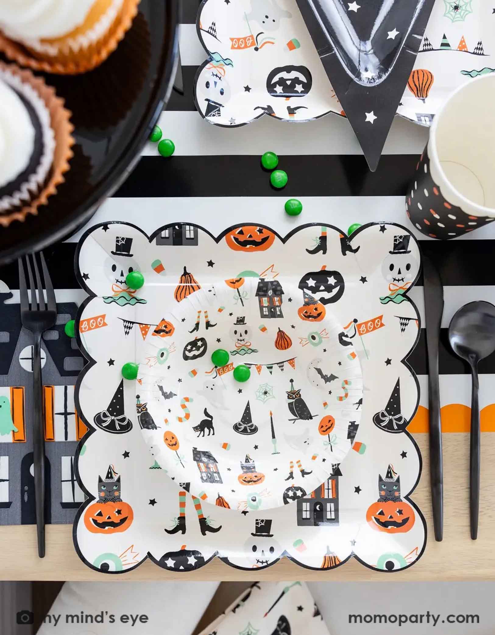 A overhead shot of a festive Halloween-themed table setting with Momopartys's Trick or Treat dinner plates and bowls, the haunted mansion shaped napkins and black cutlery set on a black orange and white striped table runner fro Momopartys. On the table there's a cauldron filled with green gum balls and some green candies spread across the table. A perfect set up for kid's friendly Halloween celebration.