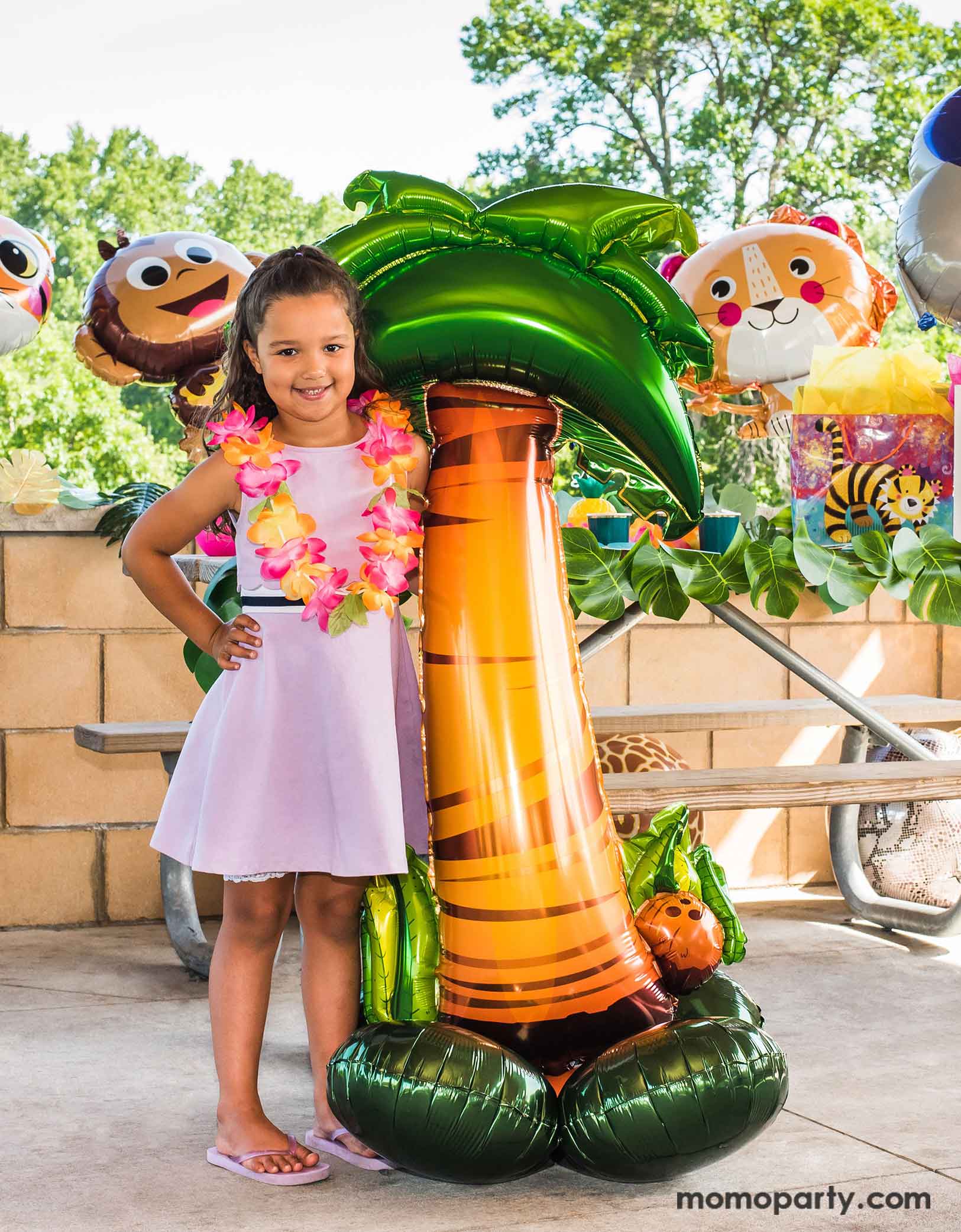 A little girl wearing Hawaiian leis stand next to Momopartys's 56" airloonz palm tree foil balloon in a tropical, luau, Hawaiian themed party decorated with animal themed foil balloons and palm tree party garland. 