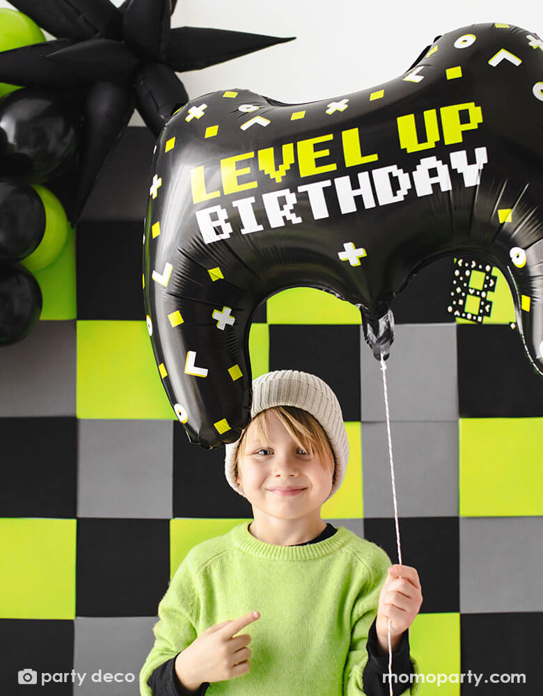 A young boy in lime green colored sweater with black long sleeves holding Momopartys's 25.5 "x 18" video controller shaped foil balloon featuring LEVEL UP BIRTHDAY message. Behind him is a video game themed party backdrop with black, grey and lime squares adorned by a balloon garland in black and lime colors and a jumbo black starburst foil balloon - a perfect decoration for a kid's video game themed birthday party.