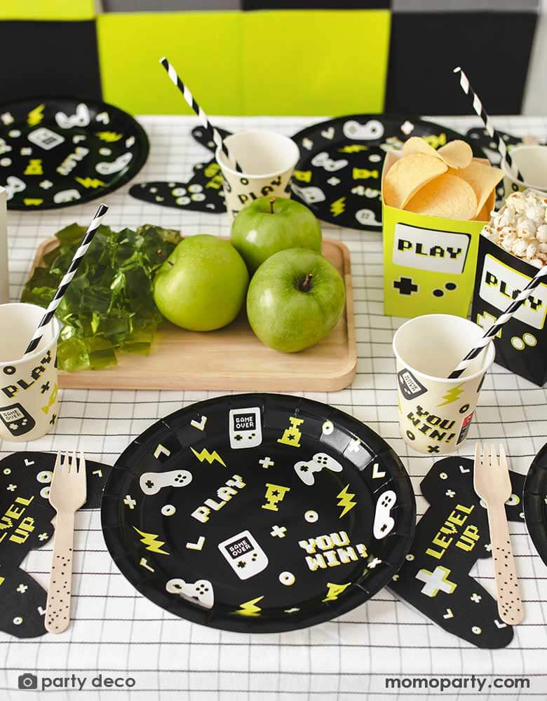 A video game themed party table featuring Momopartys's black Video Game round party plates with video game devices and controller motifs on them. Next to the plates are a game controller shaped napkins with "LEVEL UP" message on them, with the coordinating video game party cups with black striped paper straws from Momopartys. In the middle of the table, there are some green apples and green jellos next to two game boy shaped paper snack box filled with popcorn and chips.