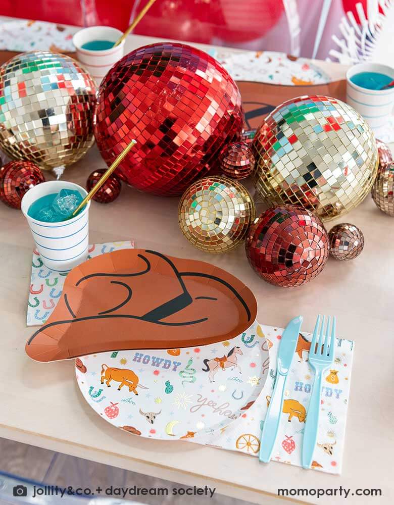 A western cowboy themed party table tablescape featuring Momopartys's cowboy themed tableware by Jollity Co. and Daydream Society including the round cowboy icon patterned round plate, the small napkin, the cowboy hat shaped plate, horseshoe napkin. With disco ball decorations in red and gold in different sizs, making this a modern yet fun tablescape for kid's rodeo, Western or cowboy themed birthday celebration. A perfect inpso for kid's "First Rodeo" first birthday celebration.  