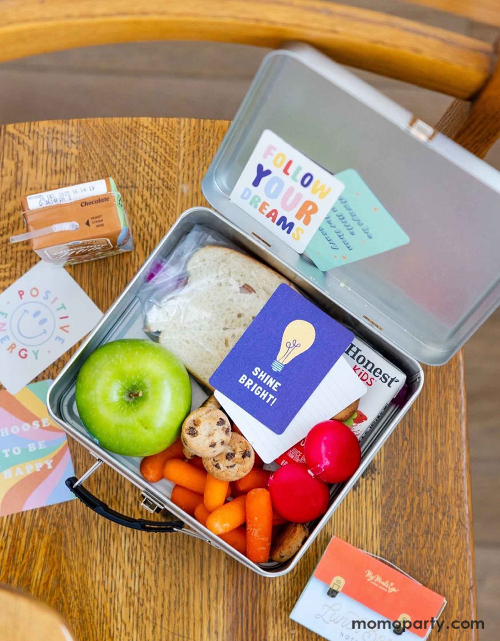 An overhead shot of a kid's lunch box on a school chair. In the lunch box includes a sandwich, an green apple, a juice box,   some cheese, veggies and cookies. There's a  card with "Shine Bright!" on it. Next to the lunch box are more lunchbox cards around and Momopartys's Pastel Lunch Box Cards package. 